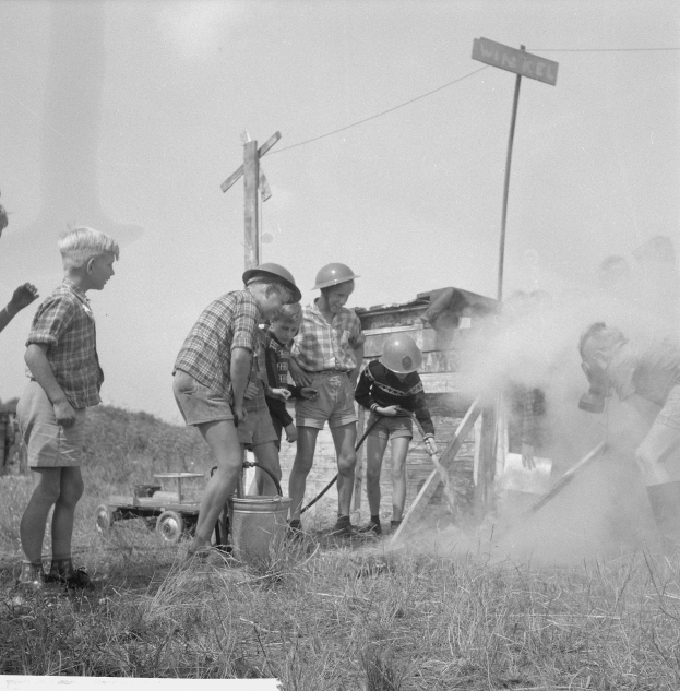 Schwarzes und wei├čes Bild von Kindern mit Helmen um einen Hydranten in einem grasigen Feld, einige halten Gegenst├Ąnde, mit Strommasten und einem Schild im Hintergrund.