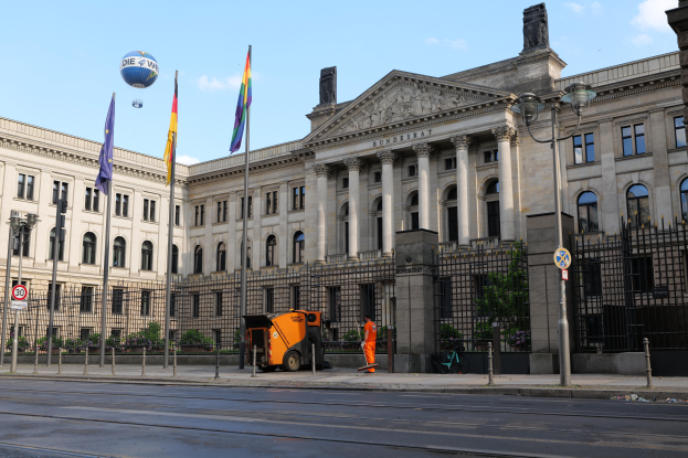 Das Reichstagsgebäude in Berlin, Deutschland, ein großes Gebäude mit zahlreichen Fenstern, umgeben von Straßenlaternen, Fahrzeugen, Menschen und Bäumen unter einem bewölkten Himmel.
