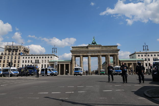 Eine Gruppe von Polizisten steht vor dem Brandenburger Tor in Berlin, Deutschland, mit seinen Säulen, der Statue und den umliegenden Gebäuden, Fahrzeugen, Laternenmasten und Verkehrszeichen unter einem bewölkten Himmel.