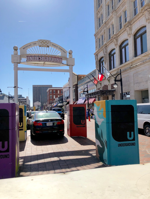 Stadtstraße mit parkenden Autos, einem dekorativen Bogen, Gebäuden, Laternen, Flaggen, einem Müllcontainer im Vordergrund und Himmel im Hintergrund.
