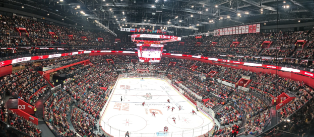 Eine große Indoor-Hockey-Halle mit Zuschauern auf den Rängen, heller Deckenbeleuchtung und einem zentralen Scoreboard-Bildschirm während eines Spiels.