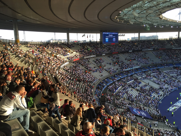 Große Zuschauermenge in einem Stadion bei einem Fußballspiel, mit einer Bühne auf der rechten Seite, Fahnen, Stangen, einem Bildschirm und dem Allianz Stadion in München im Hintergrund.