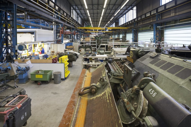 Ein großer Lagerraum voller Maschinen, darunter ein Landfahrzeug auf der rechten Seite, Metallobjekte auf der linken Seite und Lampen an der Decke.