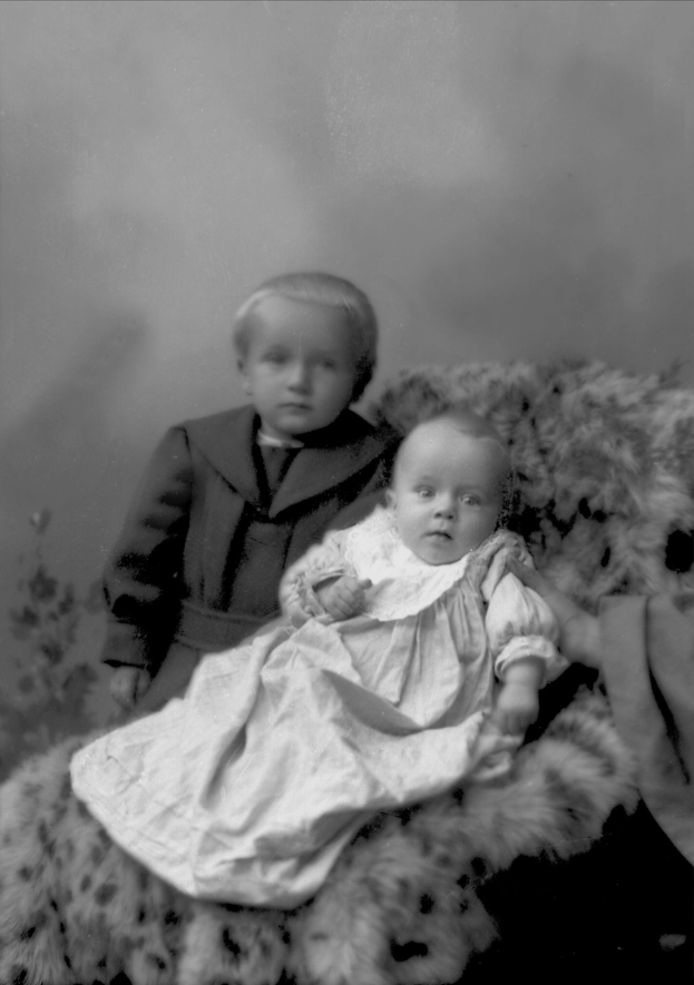 Ein altes Schwarz-Weiß-Foto eines Jungen in einem Anzug und eines Mädchens, das ein Baby hält, nebeneinander sitzend.