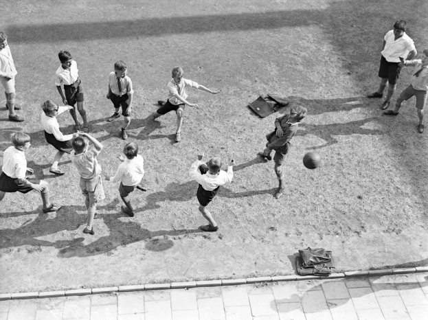 Eine Gruppe junger Jungs in Shorts und Fußballschuhen, die auf einem Feld Fußball spielen, mit Taschen, die auf dem Boden verstreut sind, in Schwarz-Weiß.