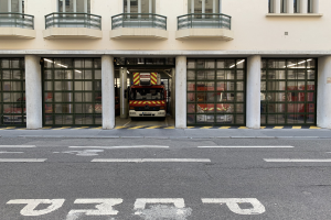 Feuerwache mit Feuerwehrauto davor, mit Fenstern, Geländern, Säulen und Glastüren und einer Straße unten.
