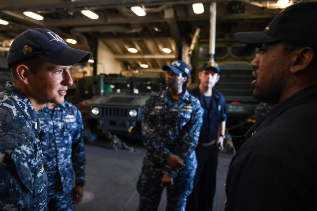 Gruppe von Männern in US-Navy-Uniformen, die vor einem Fahrzeug mit einer Wand und Deckenbeleuchtung im Hintergrund sprechen.
