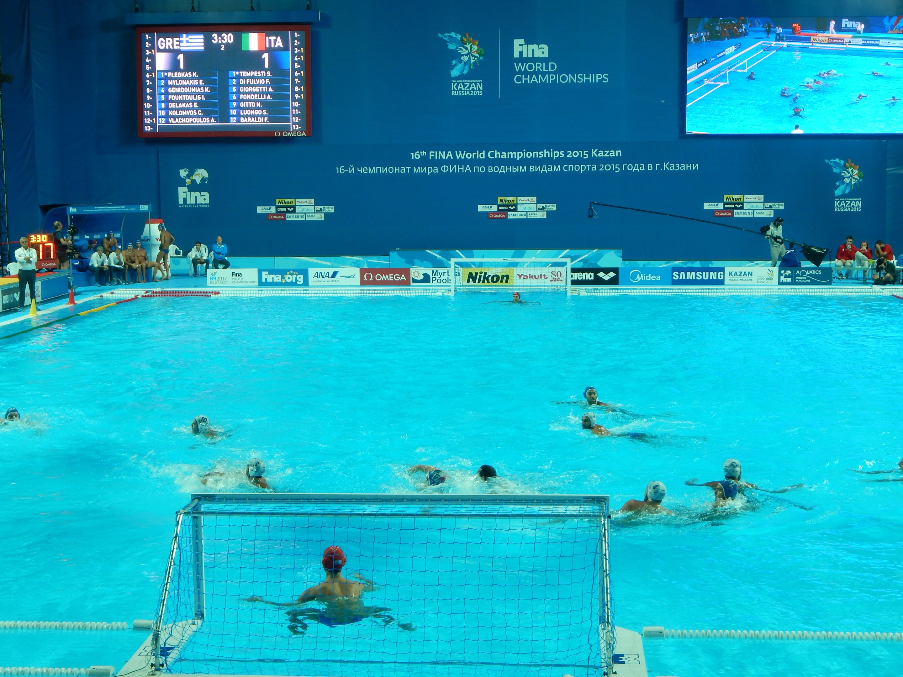 Wasserballspieler bei den 2016 FINA-Weltmeisterschaften in Kazan im Pool gegeneinander antretend, mit Zuschauern links und einem Anzeigebrett mit Event-Branding im Hintergrund.