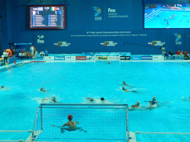 Wasserballspieler bei den 2016 FINA-Weltmeisterschaften in Kazan im Pool gegeneinander antretend, mit Zuschauern links und einem Anzeigebrett mit Event-Branding im Hintergrund.