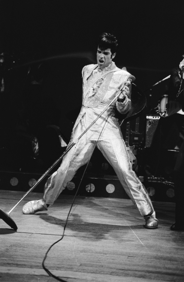 Elvis Presley in weißer Bühnenkleidung performt auf der Bühne des Apollo Theaters in Las Vegas und hält ein Mikrofon in der Hand, während ein weiterer Musiker rechts von ihm steht.