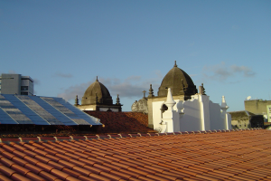 Stadtansicht mit Gebäuden im Vordergrund und einem blauen Himmel im Hintergrund, mit Solarpanelen auf einem Dach, die den Einsatz erneuerbarer Energien anzeigen.