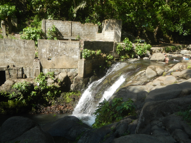 Ein kleiner Wasserfall fließt eine felsige Klippe hinunter in einem üppigen grünen Wald, mit Menschen, die im Wasser schwimmen und einer Wand im Hintergrund.