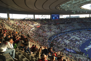 Große Menschenmenge in einem Stadion bei einem Fütballspiel mit einer Bühne, Fahnen, Masten und einem Bildschirm im Hintergrund.