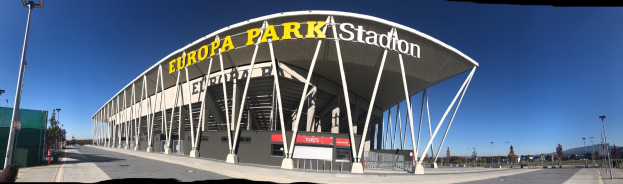 Großes Stadiongebäude mit Text, umgeben von Pfählen, Lampen, Bäumen und einem Zaun unter einem sichtbaren Himmel.