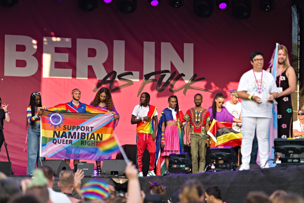 Gruppe von Menschen auf einer Bühne mit einem "Berlin as Fuck"-Schild mit Lautsprechern, Mikrofonen und einer Menge unten, die Hände hebt.