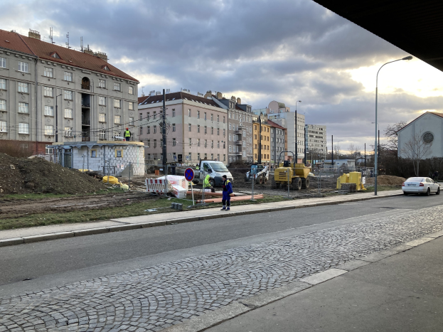 Stadtstraße mit parkenden Autos, Gebäuden, Laternen, Bäumen, einem bewölkten Himmel und einer Straßensperre, wo Menschen gehen.