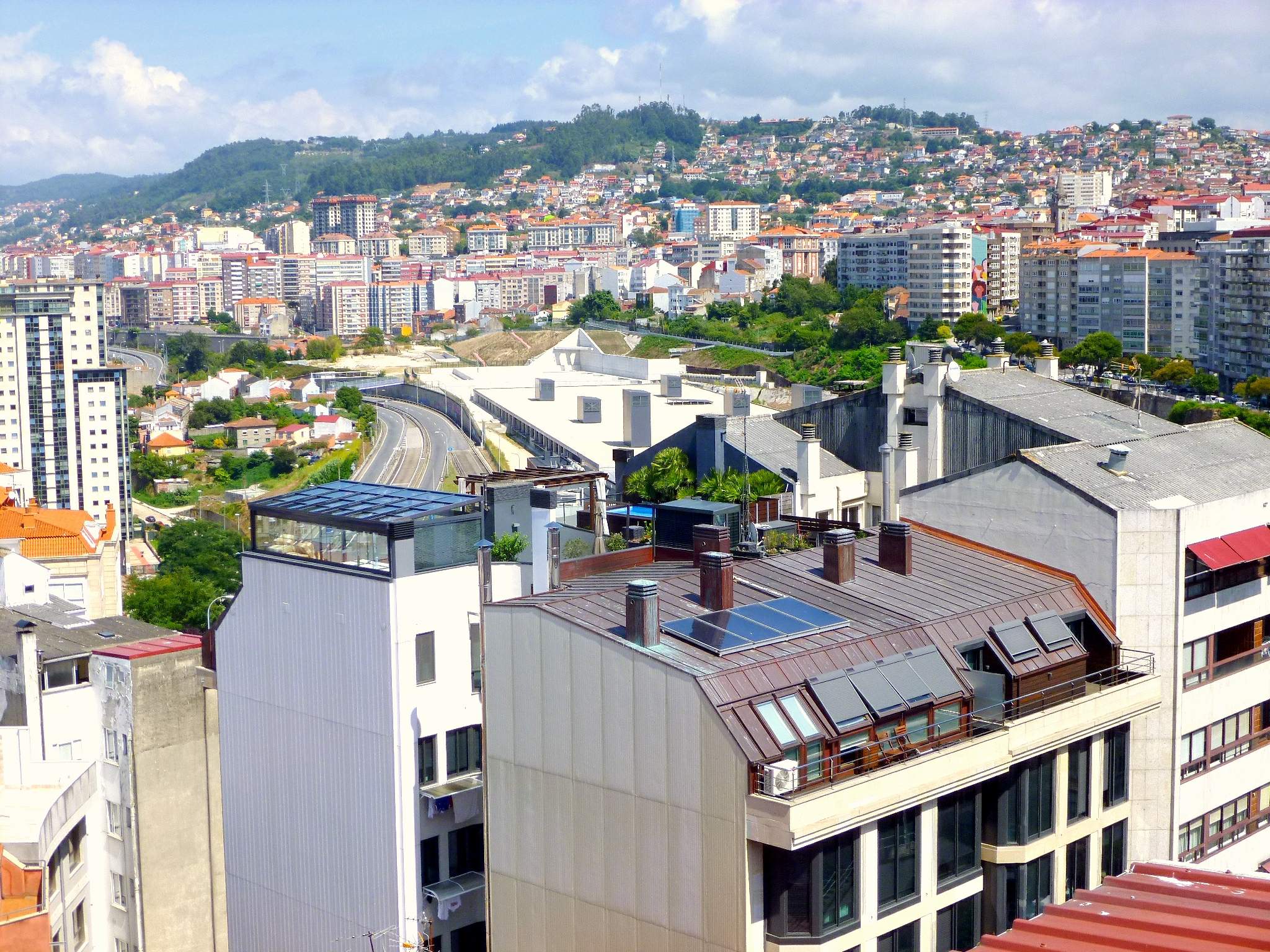 Eine Stadtansicht von einem hohen Standpunkt aus, mit zahlreichen Gebäuden, Bäumen, Straßen, Fahrzeugen und fernen Bergen unter einem bewölkten Himmel, mit Solarpanelen auf dem Dach eines Gebäudes.