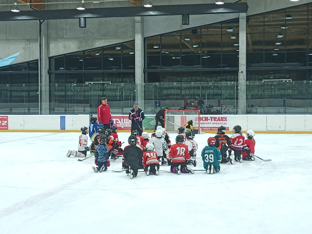 Eine Gruppe von Kindern in Helmen und mit Hockey-Schlägern sitzt auf einer Eisfläche, mit einer Wand aus Glas und Säulen, Deckenleuchten und Texttafeln im Hintergrund.