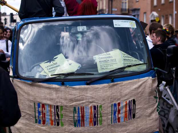 Gruppe von Menschen um ein Auto mit einer "Legalise Cannabis Ireland"-Plakette versammelt, umgeben von Gebäuden unter einem klaren blauen Himmel, mit Papieren im Auto sichtbar.