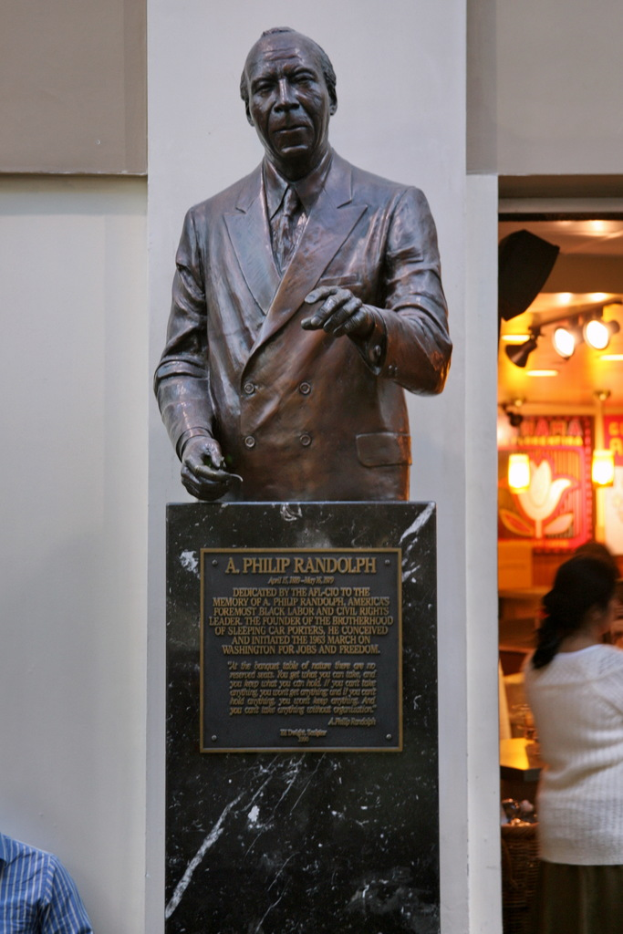 Eine Statue mit Text darunter, eine Frau rechts mit ein paar Gegenständen und eine Person links unten, alles vor einer Wand.