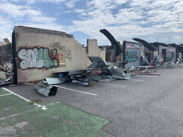 Ein Parkplatz voller Trümmer, darunter Metallschrott, mit einer links graffitibeschmierten Wand und beschädigten Gebäuden im Hintergrund, die die Auswirkungen von Hurrikan Katrina zeigen.