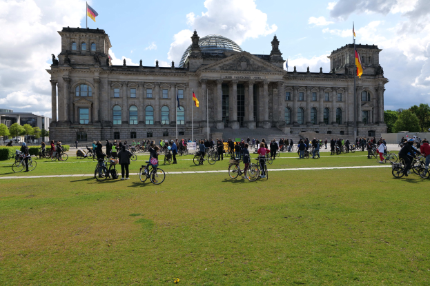 Eine Gruppe von Menschen, die vor dem Reichstaggebäude in Berlin, Deutschland, Fahrräder fahren, mit sichtbaren Fenstern, Säulen und Fahnenmasten des Gebäudes, umgeben von Grün unter einem bewölkten Himmel.