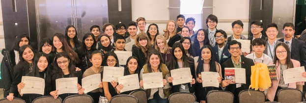 Eine Gruppe von Menschen hält Zertifikate in der Hand und posiert für ein Foto bei einer Abschlussfeier mit Stühlen und einer Wand im Hintergrund.
