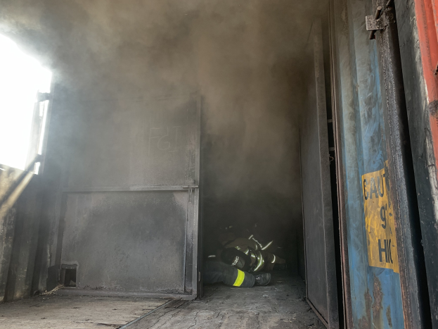 Feuerwehrmann in voller Montur vor einer raucherfüllten Tür eines brennenden Gebäudes mit einer Tür mit der Beschriftung "BX1" auf der rechten Seite und einem undeutlichen Objekt auf der linken Seite.