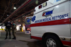 Ein Rettungswagen der American Medical Response (AMR) steht in einem Gebäude, mit zwei maskierten Personen davor; links ein Fahrzeug und weitere Personen, Lichter und Rohre im Hintergrund.