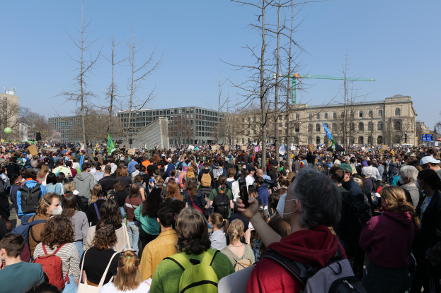 Eine große Menschenmenge mit Schildern und Taschen steht vor einem Gebäude mit Fenstern, umgeben von Bäumen und einem klaren Himmel, was auf eine Klimaprotest vermuten lässt.