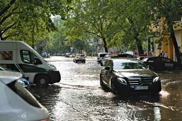 Eine überflutete Straße in Berlin, Deutschland, mit Autos, die durch das Wasser fahren, Menschen, die auf dem Gehweg gehen, Bäume, die die Straße säumen, Gebäude im Hintergrund und Schilder und Pfosten, die teilweise untergetaucht sind.