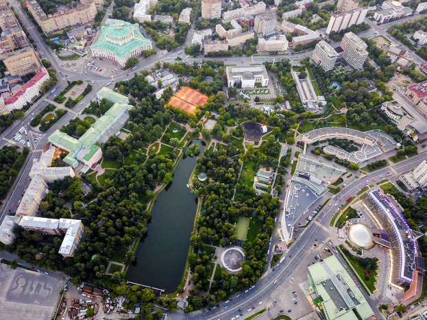 Luftaufnahme einer Stadt mit dichtem Baumbestand, Straßen, Fahrzeugen und Grünflächen, mit einem zentralen Park inmitten von Bäumen und Vegetation.
