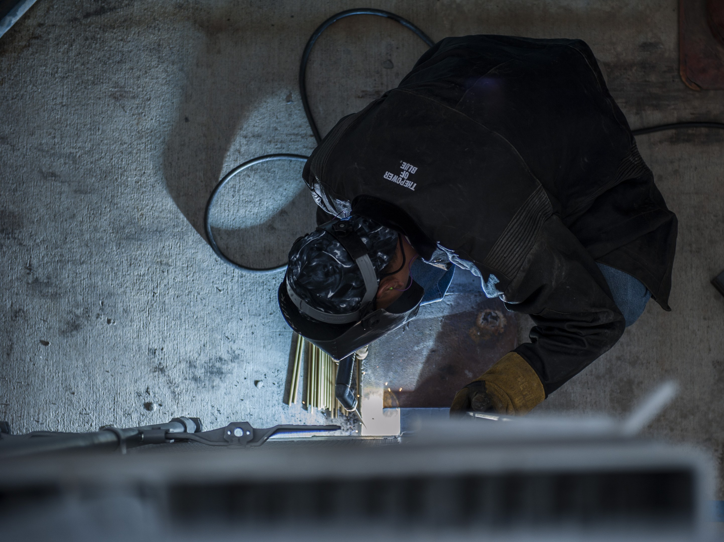 Ein Schweißer in Schutzausrüstung verwendet eine Schweißmaschine an einem Metallstück, mit Werkzeugen und Gegenständen, die auf dem Boden verstreut sind.