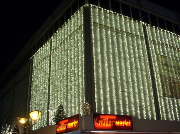 Ein Gebäude in Berlin, Deutschland, geschmückt mit Weihnachtslichtern, umgeben von Bäumen und Laternenmasten, mit Texttafeln in der Nähe und einem sichtbaren Himmel im Hintergrund.