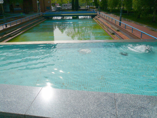 Großer rechteckiger Schwimmbad im Park, umgeben von Zaun und Laternenmasten, mit Bäumen und parkenden Fahrzeugen drumherum, vor Buildings und einem klaren blauen Himmel.