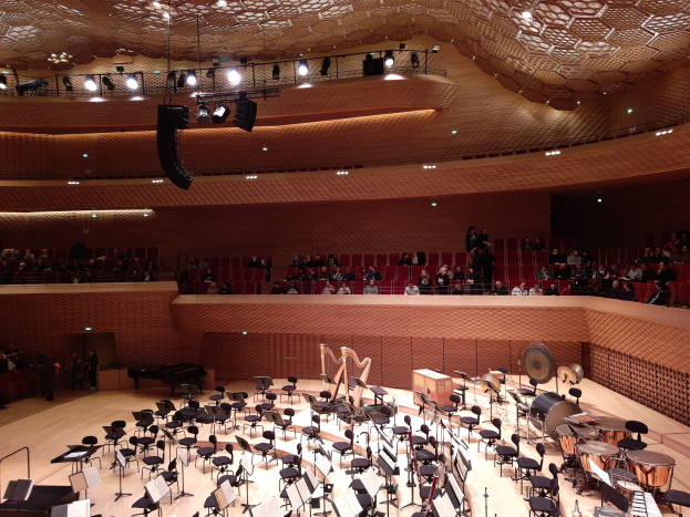 Ein großer Saal voller Musikinstrumente, Stühle, Ständer mit Papieren und Menschen mit beleuchteter Decke, repräsentiert den Hauptsaal der Berliner Philharmonie.