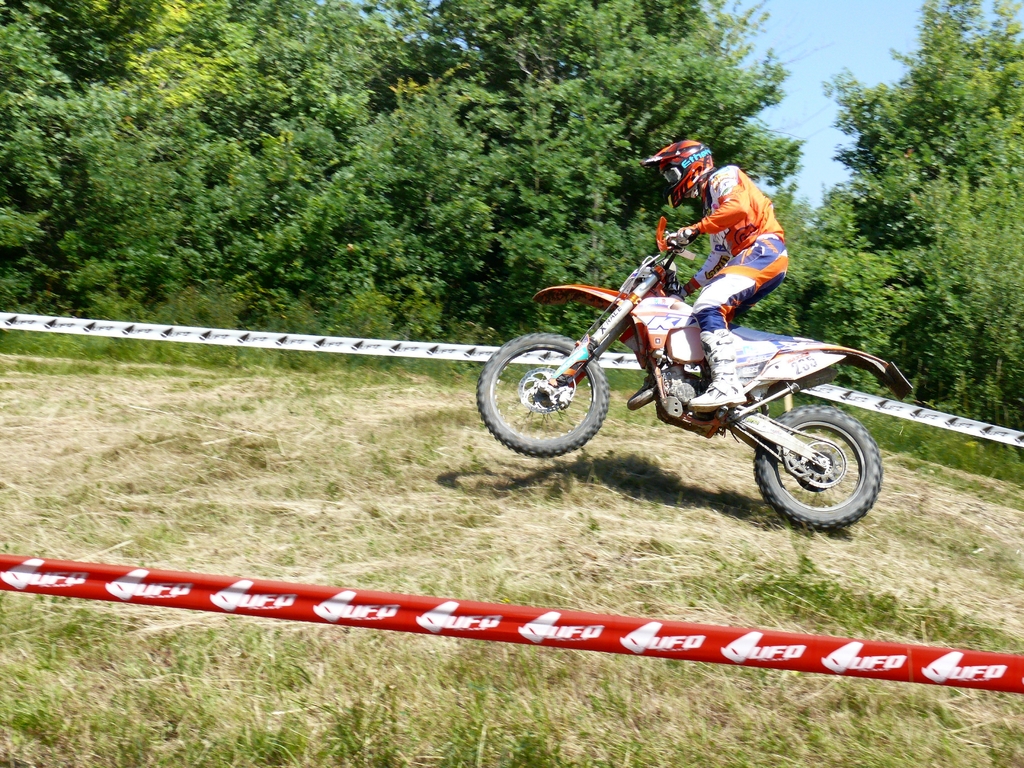 Eine Person fährt Fahrrad und trägt einen Helm, umgeben von Absperrband, mit Bäumen, Gras und Himmel im Hintergrund.