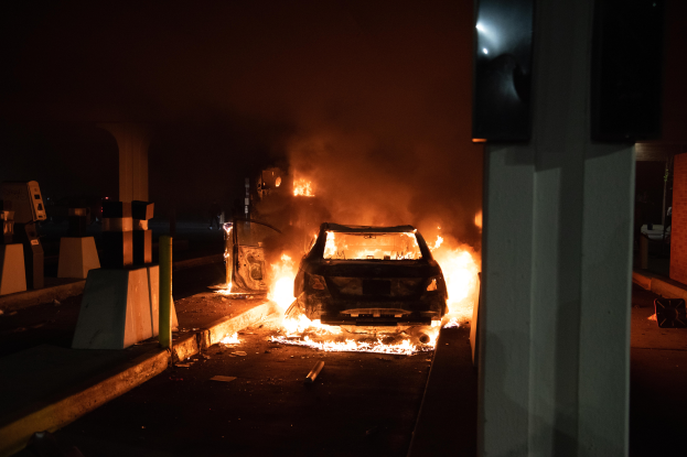 Ein Auto steht in Flammen am Straßenrand bei Nacht, mit einer Säule rechts und ein paar Gegenständen links, was auf einen Tankstellenbrand im Hintergrund hinweist.