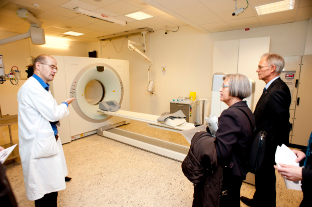 Ein Arzt in weißer Uniform erklärt eine CT-Scan-Maschine zwei Menschen in schwarzen Mänteln, die aufmerksam zuhören.