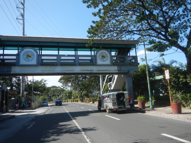 Ein Bus fährt eine von Bäumen gesäumte Straße mit einer Brücke im Hintergrund, mit Text und Logos auf Schildern, Strommasten mit Drähten, Laternenmasten und einigen Fußgängern auf dem Gehweg unter einem bewölkten Himmel.