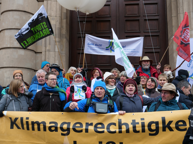 Eine Gruppe von Menschen vor einem Gebäude mit Säulen und einer Tür, die ein Transparent mit Text, Fahnen mit Stangen und Luftballons halten, an einer Klimademonstration in Deutschland teilnehmend.