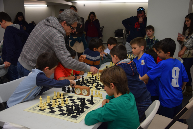 Kinder spielen beim Schach-Wettbewerb, während Erwachsene von Hintergrund und Seiten zuschauen.