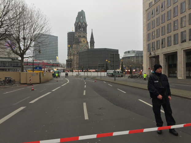 Ein Polizeibeamter in schwarzer Uniform und Mütze steht auf einer Straße in Berlin umgeben von Absperrbaken, Fahrrädern, Laternenpfählen, Bäumen und Gebäuden, mit einem Uhrenturm und einem klaren blauen Himmel im Hintergrund.