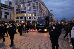 Eine Gruppe von Menschen steht in der Nähe eines Lastwagens auf einer Straße, die von Gebäuden, Bäumen und Laternen gesäumt ist, unter einem bewölkten Himmel, einige tragen Mützen und Masken, mit einem Band an einem Pfahl im Vordergrund.
