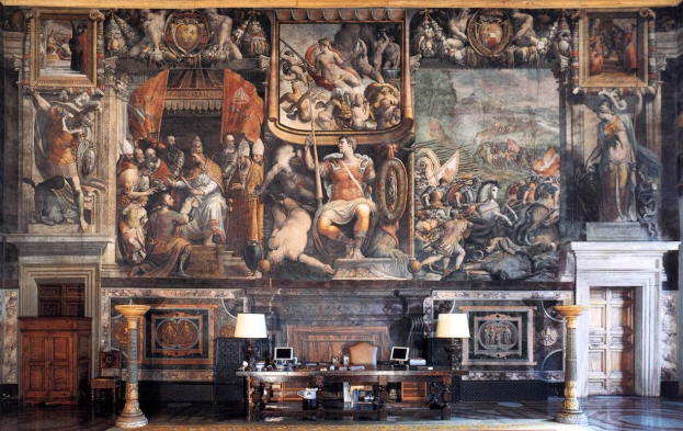 Ein prächtiger Raum im Palazzo della Signoria in Florenz, Italien, mit einem großen Gemälde an der Wand, einem Tisch mit Gegenständen, zwei Lampen, zwei Türen und einem Teppich.