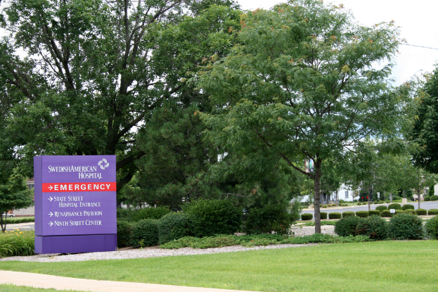 Ein grasbewachsener Bereich mit einem violetten Notfallschild, auf dem "NOTFALL" in fetter Schrift steht, umgeben von Bäumen, Sträuchern, Gebäuden und einer Straße im Hintergrund.