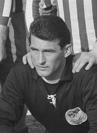 Schwarzes und weißes Foto eines Mannes, der auf einem grünen Fussballfeld sitzt und ein T-Shirt und eine Kette trägt, mit zwei Personen hinter ihm.