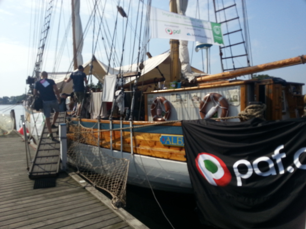 Ein Segelschiff auf der rechten Seite, eine hölzerne Brücke auf der linken Seite mit zwei Männern, die das Schiff mit einem hölzernen Block betreten, unter einem bewölkten Himmel, eingerahmt von einem Banner oben.