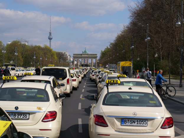 Eine lange Reihe von Taxis, die entlang einer belebten Straße in Berlin, Deutschland, geparkt sind, mit Fahrradfahrern und Fußgängern auf dem Gehweg, Laternenpfählen und Bäumen, die die Straße säumen, und Gebäuden, einem Bogen und einem Turm im Hintergrund unter einem bewölkten Himmel.