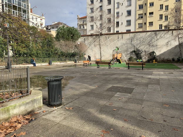 Ein Park mit Bänken, einem Mülleimer, trockenen Blättern auf dem Boden, einem Metallzaun, Bäumen, Pflanzen, einer Wand, Pfählen, einem Straßenpfahl, Menschen auf den Bänken sitzend, Kindern auf der Wiese spielend, Gebäuden mit Fenstern und einem bewölkten Himmel.